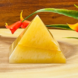 May include: A yellow crystal pyramid on a wooden surface. The pyramid is smooth and has a light yellow color. It is surrounded by green leaves and orange flowers.