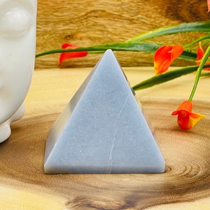 May include: A light blue crystal pyramid, with a smooth, polished surface. The pyramid is sitting on a wooden surface.
