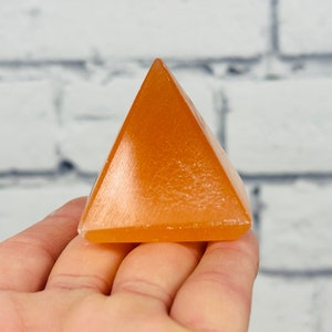 May include: A small, orange, crystal pyramid. The pyramid is smooth and polished, with a slight sheen.