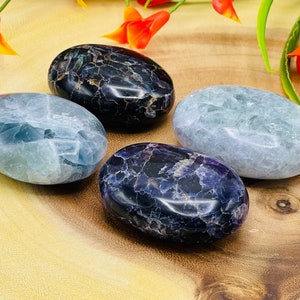 May include: Four smooth, polished, oval-shaped stones in shades of blue, purple, and black. The stones are arranged on a wooden surface.