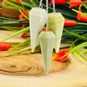 May include: Three crystal pendulums, one white and two green, hanging from silver chains. The pendulums are on a wooden surface with red flowers in the background.