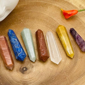 May include: Seven chakra crystal wands in a rainbow of colors, including red, orange, yellow, green, blue, indigo, and violet. The wands are arranged in a line on a wooden surface.