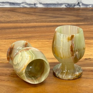 May include: Three green and brown onyx wine glasses. Two glasses are lying on their sides, while the third stands upright. The glasses have a unique, natural pattern and are set against a wooden surface.
