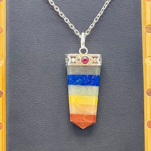 May include: A silver chain necklace with a seven chakra pendant. The pendant is made of layered stones in red, orange, yellow, green, blue, indigo, and violet. A small red gemstone is set in the silver bezel.