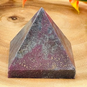 May include: A purple and grey pyramid-shaped crystal with gold flecks. The crystal is sitting on a wooden surface.