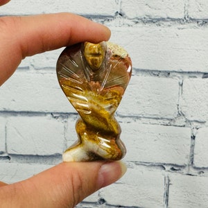 May include: A small, hand-carved stone sculpture of a cobra in a coiled position. The sculpture is made of a light brown and white stone with brown and orange markings.