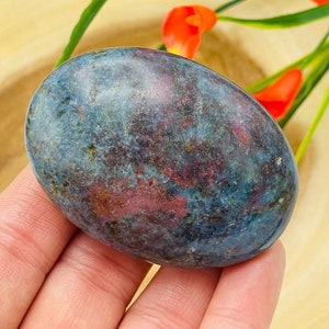 May include: An oval-shaped stone with a mottled appearance, featuring a blend of blue, red, and green hues. The stone is held in a hand, with a wooden surface and red flowers in the background. The stone's surface has a polished look.