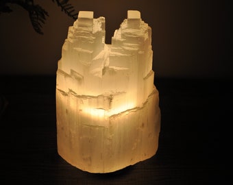 May include: A white selenite crystal lamp with a warm glow emanating from the base. The crystal is naturally formed with a rough, textured surface.
