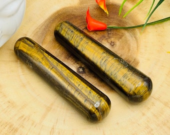 May include: Two tiger's eye crystal wands, one lying on top of the other, on a wooden surface. The wands are a golden brown color with dark brown stripes.