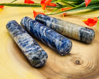 Puede incluir: Tres varitas cilíndricas de cristal de sodalita con extremos redondeados. Las varitas muestran una mezcla de tonos azules, blancos y grises. Los cristales están dispuestos sobre una superficie de madera, con acentos florales rojos y verdes.