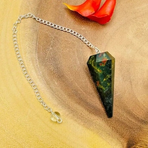 May include: A green, red, and black crystal pendulum on a silver chain. The pendulum is a point shape and is hanging from a wooden surface.