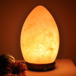 May include: A warm, glowing salt lamp shaped like an egg with a wooden base. The lamp is made of natural Himalayan salt crystals and emits a soft, orange light.