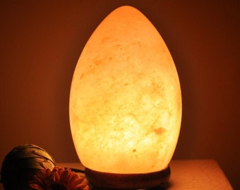 May include: A warm, glowing salt lamp shaped like an egg with a wooden base. The lamp is made of natural Himalayan salt crystals and emits a soft, orange light.
