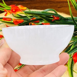 May include: A white, translucent bowl with a rounded shape and a pedestal base. The bowl is held in a hand, with green and orange flowers in the background. The bowl appears to be made of a smooth, polished material.