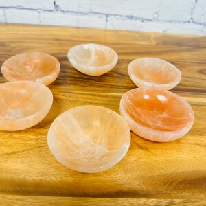 May include: Six small, round, orange-coloured bowls made of polished selenite crystal. The bowls are arranged in a circle on a wooden surface.