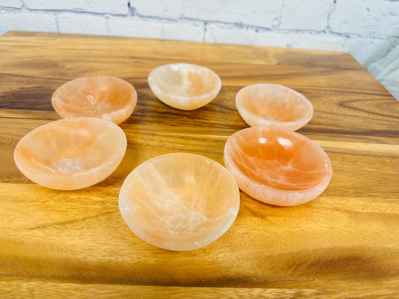 May include: Six small, round, orange-coloured bowls made of polished selenite crystal. The bowls are arranged in a circle on a wooden surface.