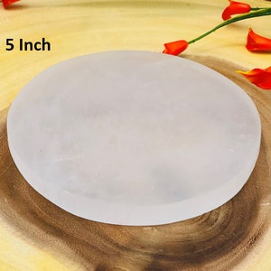 May include: A 5 inch white selenite charging plate, a smooth, round, white disc on a wooden surface.