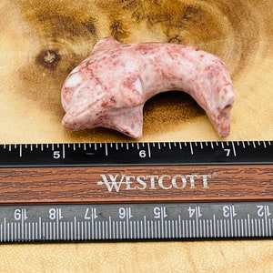 Puede incluir: Una figurita de delf&iacute;n de m&aacute;rmol rosa y rojo tallada a mano. El delf&iacute;n mide aproximadamente 13 cm de largo y est&aacute; sentado sobre una superficie de madera.