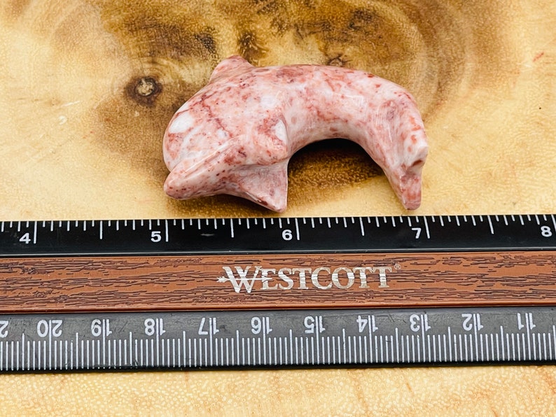 Puede incluir: Una figurita de delf&iacute;n de m&aacute;rmol rosa y rojo tallada a mano. El delf&iacute;n mide aproximadamente 13 cm de largo y est&aacute; sentado sobre una superficie de madera.