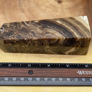 May include: A brown and cream-colored crystal obelisk with a layered, swirling pattern. The obelisk is rectangular with a slanted top. A ruler with inch measurements is in the foreground.