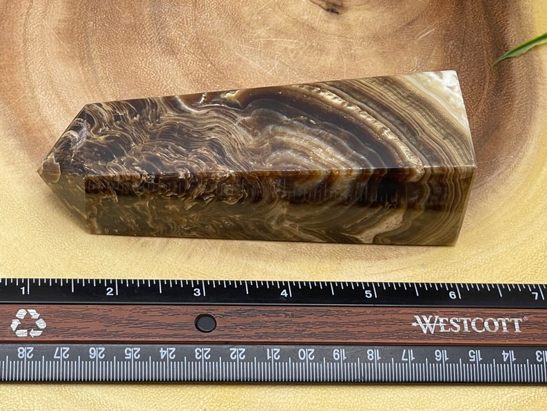 May include: A brown and cream-colored crystal obelisk with a layered, swirling pattern. The obelisk is rectangular with a slanted top. A ruler with inch measurements is in the foreground.