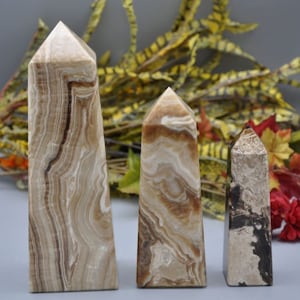 May include: Three stone obelisks with a polished finish. The tallest is approximately 8 inches tall, featuring brown and cream-colored banding. The other two are smaller, with similar color patterns and a pointed top. The stones are displayed against a blurred background.