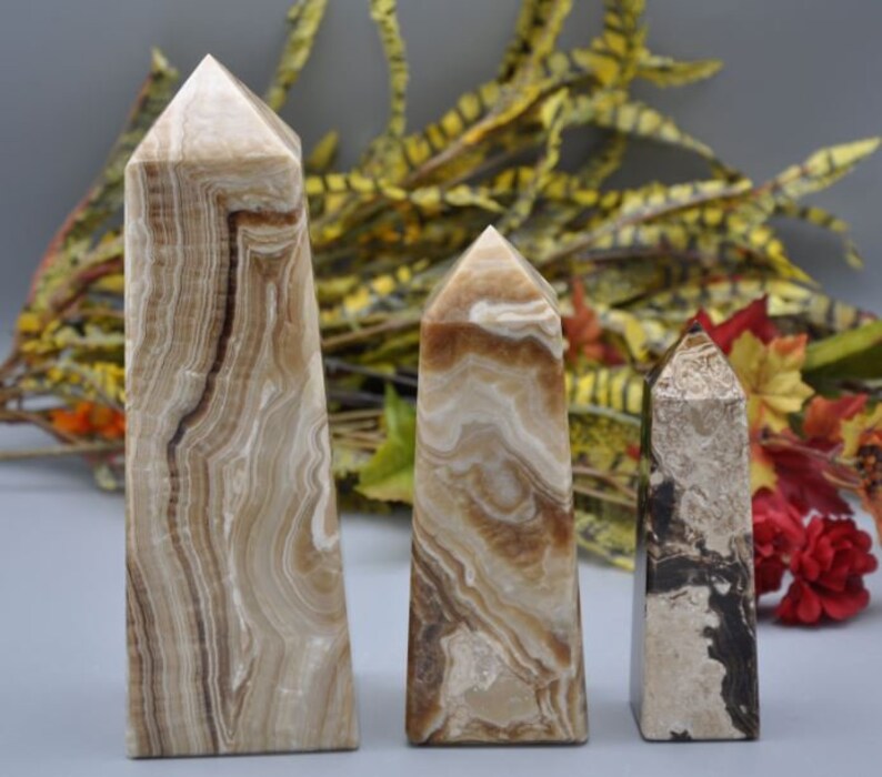 May include: Three stone obelisks with a polished finish. The tallest is approximately 8 inches tall, featuring brown and cream-colored banding. The other two are smaller, with similar color patterns and a pointed top. The stones are displayed against a blurred background.