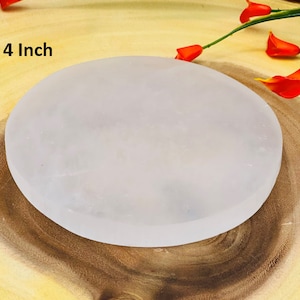 May include: A 4 inch white selenite charging plate. The plate is round and smooth, and it is sitting on a wooden surface.