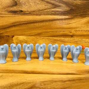 May include: A row of ten small, gray angel figurines made of polished stone. The angels are arranged in a line on a wooden surface.