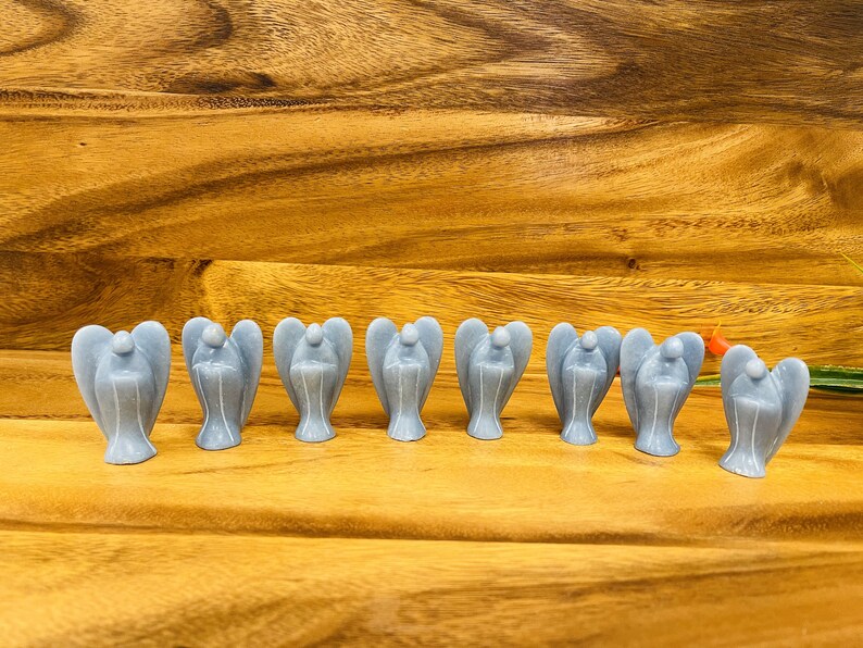 May include: A row of ten small, gray angel figurines made of polished stone. The angels are arranged in a line on a wooden surface.