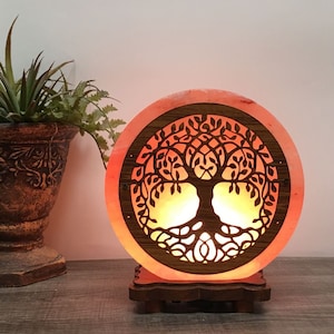 May include: A round Himalayan salt lamp with a carved tree of life design. The lamp is lit and glows orange. It is sitting on a wooden base.