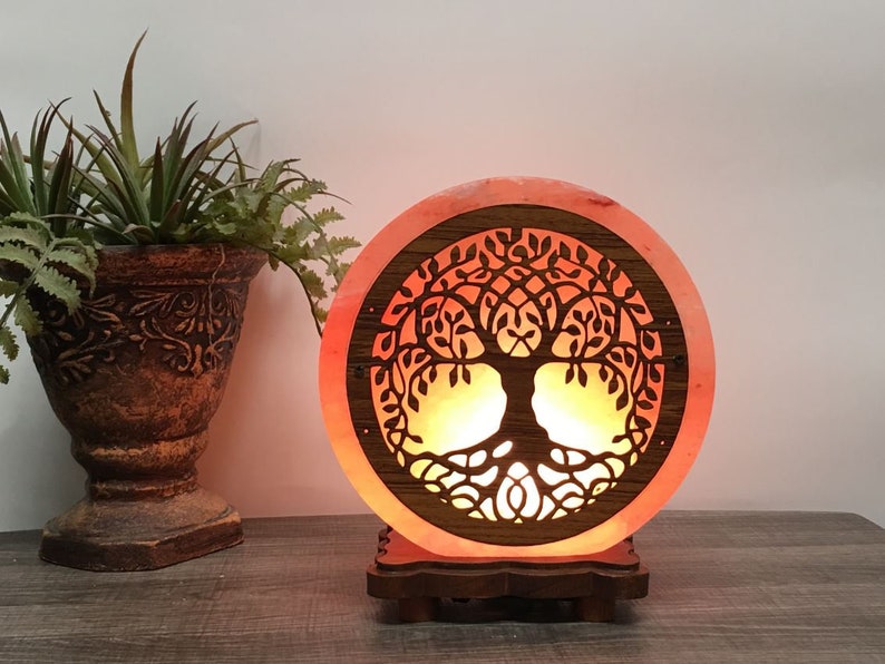 May include: A round Himalayan salt lamp with a carved tree of life design. The lamp is lit and glows orange. It is sitting on a wooden base.