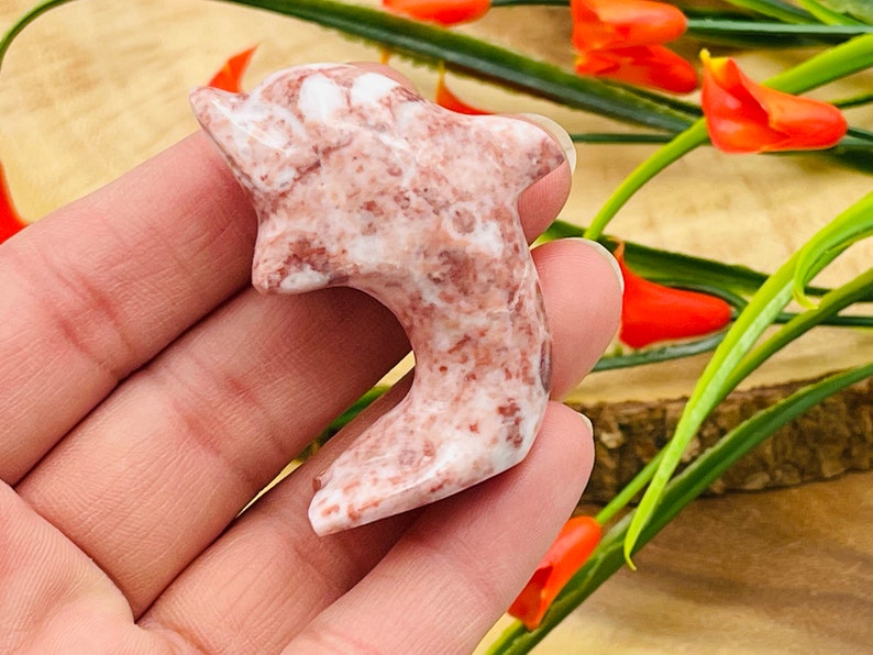 Puede incluir: Una mano sosteniendo una figurita de delf&iacute;n tallada en rosa y blanco. El delf&iacute;n est&aacute; hecho de una piedra marmoleada con un acabado liso y pulido.