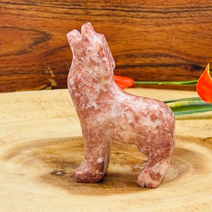 May include: A carved pink and white marble wolf figurine howling at the moon. The figurine is standing on a wooden surface.