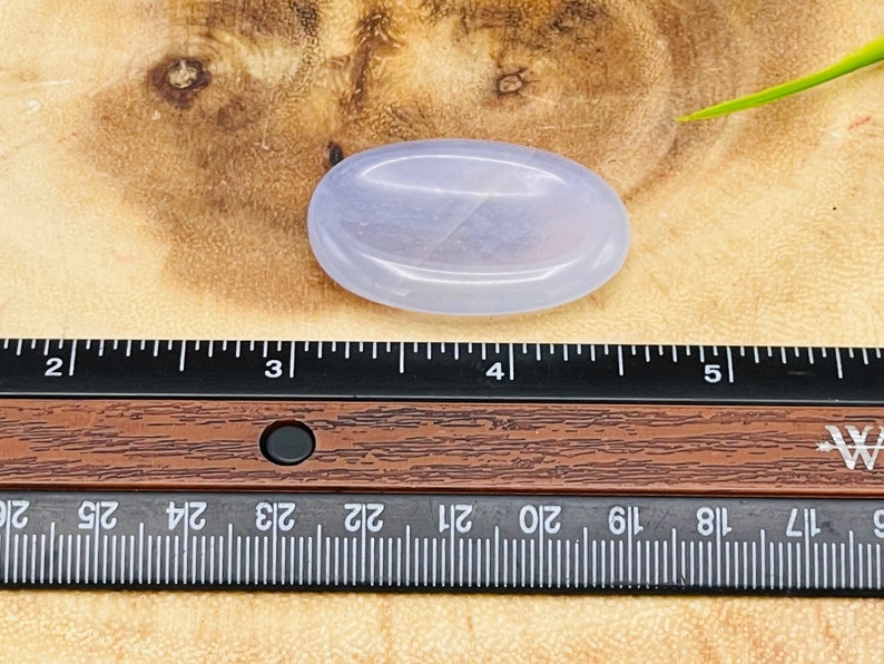 May include: A pale lavender oval-shaped polished stone, approximately 4 inches long, resting on a wooden surface.