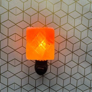 May include: An illuminated orange Himalayan salt night light. The salt lamp is rectangular with a diamond pattern and sits on a black plug. The background is a geometric pattern of white and gold lines.
