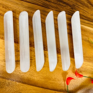 May include: Six white selenite wands, each with a rounded end, are arranged in a row on a wooden surface. The wands are smooth and polished, with a natural, crystalline texture.