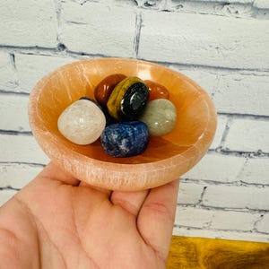 May include: A small, orange bowl made of selenite with a variety of colourful tumbled stones inside. The stones include blue, green, red, and brown.