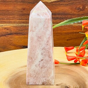 Peut inclure: Un ob&eacute;lisque en cristal rose et blanc avec un sommet pointu. L'ob&eacute;lisque est pos&eacute; sur une surface en bois.