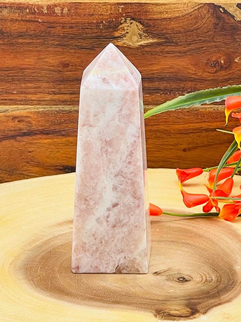 Peut inclure: Un ob&eacute;lisque en cristal rose et blanc avec un sommet pointu. L'ob&eacute;lisque est pos&eacute; sur une surface en bois.