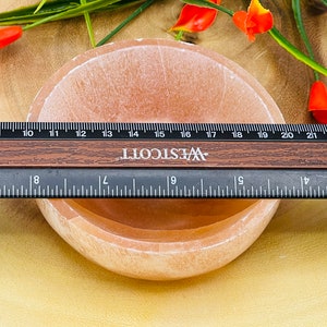 May include: A light pink, round, shallow bowl made of alabaster. The bowl is approximately 10 cm in diameter and is sitting on a wooden surface. A ruler is visible in the image, with the bowl measuring 10 cm across.