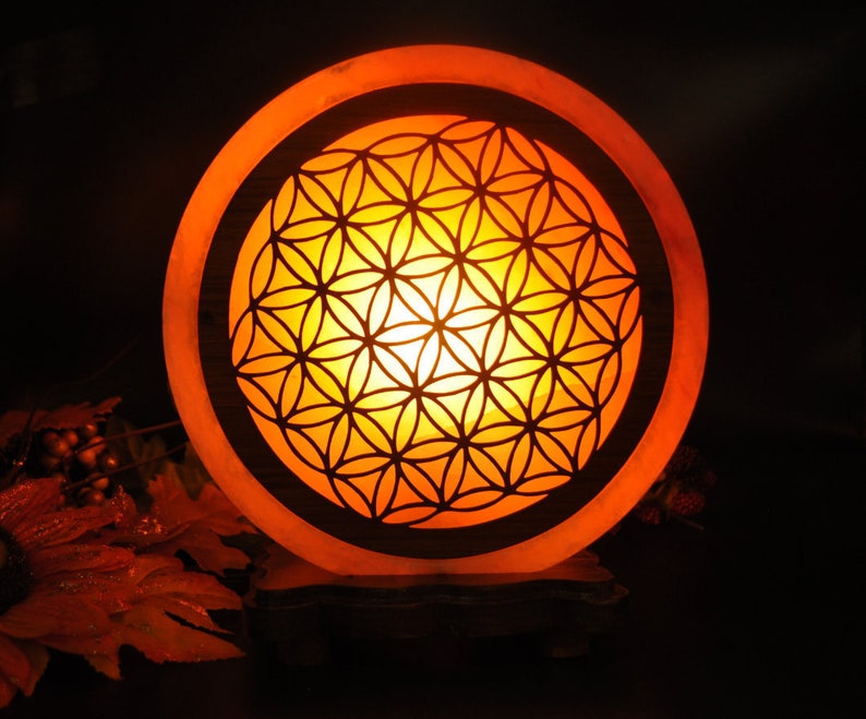 May include: A round salt lamp with a carved flower of life design. The lamp is lit and glows orange. The lamp is on a wooden base.