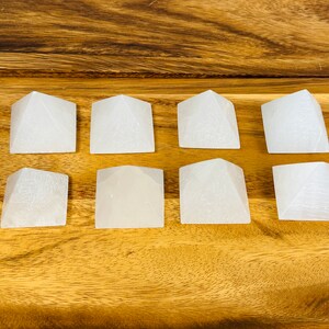Puede incluir: Ocho pir&aacute;mides de cristal de selenita blanca dispuestas en tres filas sobre una superficie de madera.