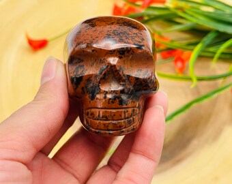 以下が含まれることがあります： 茶色と黒の色合いの彫刻されたスカルフィギュア。 スカルは手で持たれており、眼窩、鼻腔、歯のある顎の線など、その詳細な特徴が強調されています。 背景には緑と木の表面が含まれています。