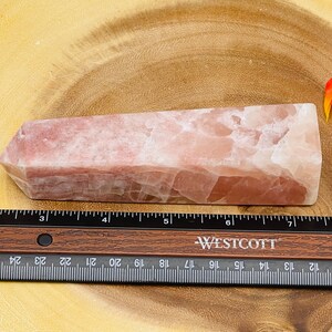 Peut inclure: Une pointe de cristal rose et blanche, d'environ 15 cm de long, est plac&eacute;e sur une surface en bois. Une r&egrave;gle est plac&eacute;e sous la pointe de cristal.