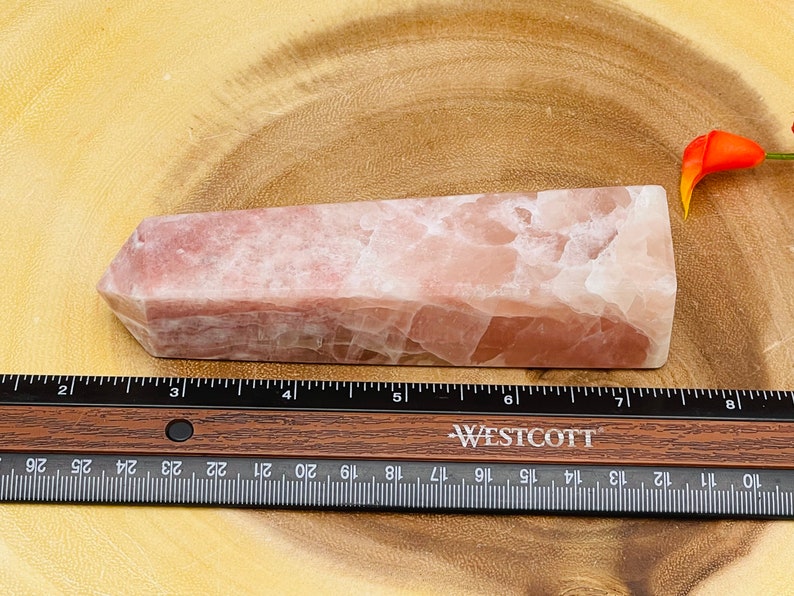 Peut inclure: Une pointe de cristal rose et blanche, d'environ 15 cm de long, est plac&eacute;e sur une surface en bois. Une r&egrave;gle est plac&eacute;e sous la pointe de cristal.