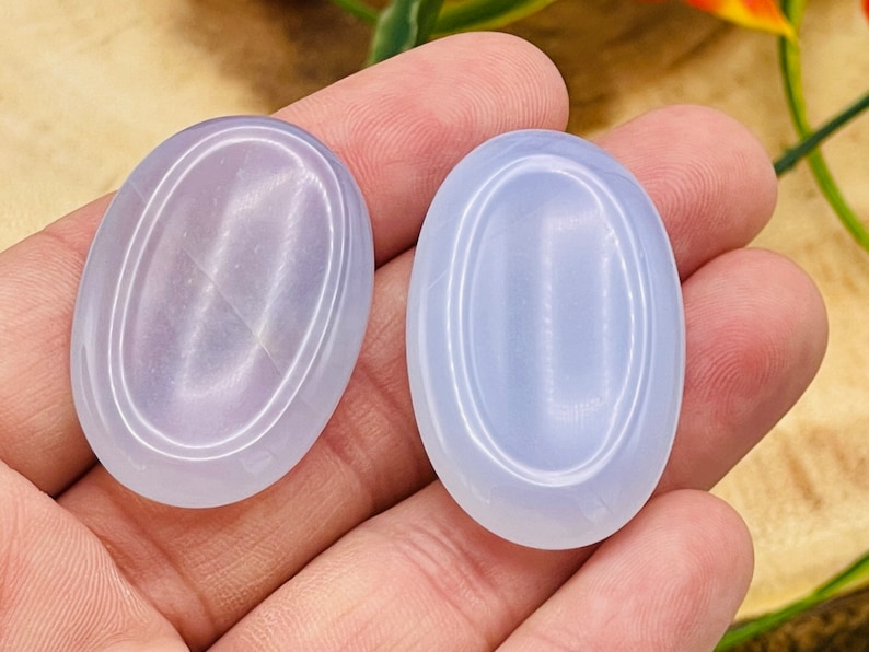 May include: Two oval-shaped, light blue, polished stones with a smooth, shiny surface. The stones have a white, concentric circle pattern.