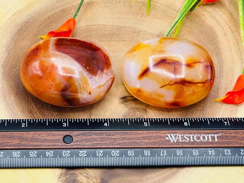 May include: Two carnelian tumbled stones, one with a white and orange swirl pattern and the other with a white and brown swirl pattern, are displayed on a wooden surface with a ruler for size reference.