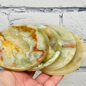 May include: Four round coasters made of natural onyx stone. The coasters have a polished finish and display a mix of green, yellow, and brown hues. The stone's natural patterns create a unique look for each coaster.