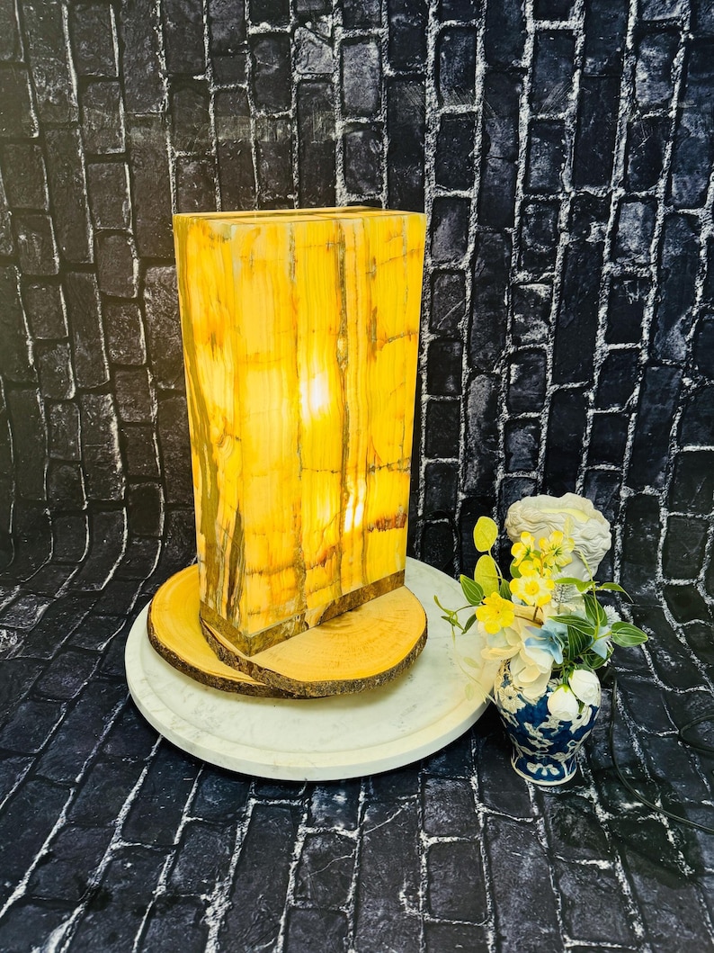 May include: A rectangular, yellow and brown onyx lamp with a wood base. The lamp is sitting on a white marble tray with two wooden rounds underneath. The lamp is lit and casting a warm glow.
